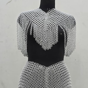 Puede incluir: Una prenda de cota de malla plateada exhibida en un maniquí negro. La cota de malla cubre los hombros, el pecho y la cintura, con una parte inferior en forma de falda. El diseño presenta un detalle de flecos en el pecho. El maniquí está contra un fondo liso.