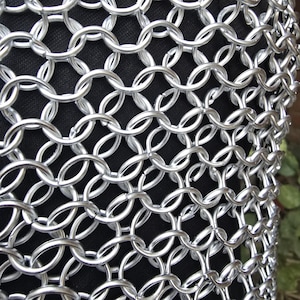 Puede incluir: Primer plano de una secci&oacute;n de cota de malla. Los anillos de metal plateado est&aacute;n entrelazados en un patr&oacute;n denso, creando una tela flexible y protectora. El fondo es un material oscuro y texturizado.