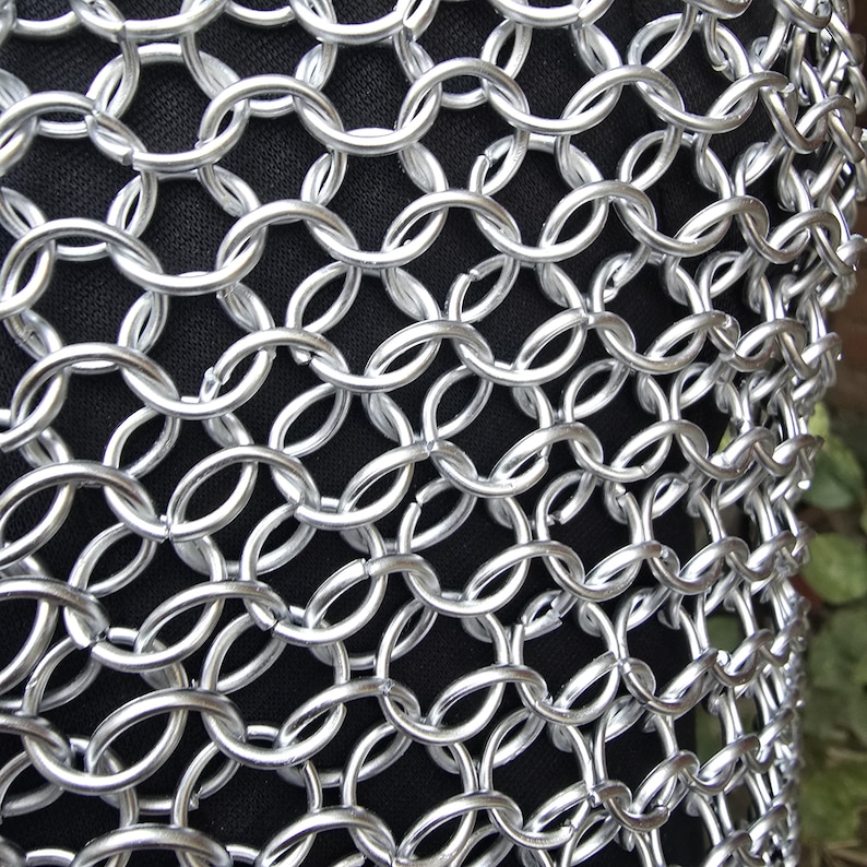 Puede incluir: Primer plano de una secci&oacute;n de cota de malla. Los anillos de metal plateado est&aacute;n entrelazados en un patr&oacute;n denso, creando una tela flexible y protectora. El fondo es un material oscuro y texturizado.