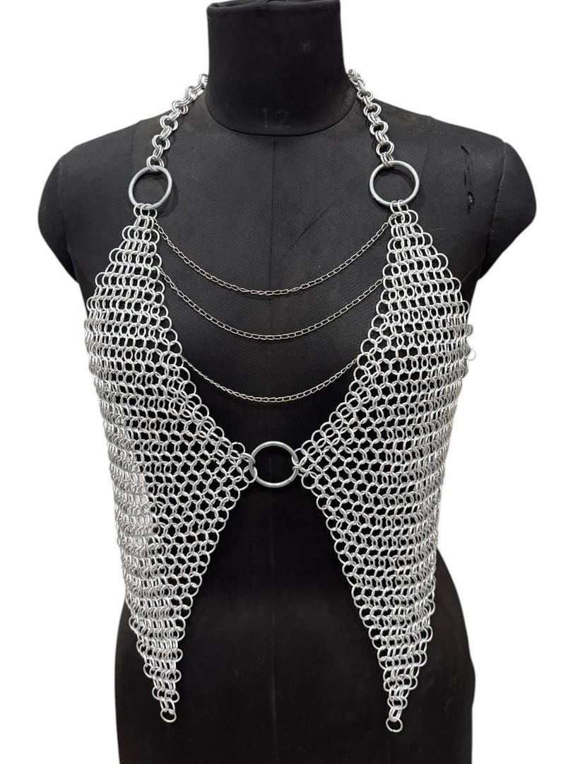 Puede incluir: Top de cota de malla plateada con dise&ntilde;o de cuello halter. La parte superior presenta una serie de anillos met&aacute;licos conectados que forman un patr&oacute;n similar a una malla. El dise&ntilde;o incluye anillos grandes en los hombros y un anillo central en la cintura, con cadenas delicadas.