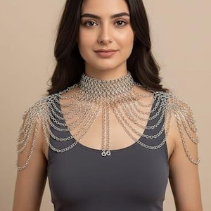 Op de afbeelding: Een zilverkleurige maliënkolder schouderketting met een hoge choker-halsband verbonden aan meerdere gedrapeerde kettinglagen die over de schouders en de borst vallen, afgewerkt met twee bungelende kettingstrengen aan de voorzijde.
