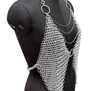 Puede incluir: Top de cota de malla plateada con un panel frontal en forma de diamante y correas de cadena. Se muestra en un maniqu&iacute; negro. La cota de malla est&aacute; hecha de peque&ntilde;os anillos met&aacute;licos entrelazados, creando una superficie flexible y texturizada. El dise&ntilde;o incluye grandes anillos circulares y cadenas delicadas.