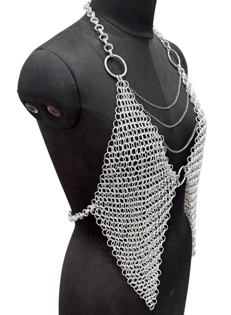 Puede incluir: Top de cota de malla plateada con un panel frontal en forma de diamante y correas de cadena. Se muestra en un maniqu&iacute; negro. La cota de malla est&aacute; hecha de peque&ntilde;os anillos met&aacute;licos entrelazados, creando una superficie flexible y texturizada. El dise&ntilde;o incluye grandes anillos circulares y cadenas delicadas.