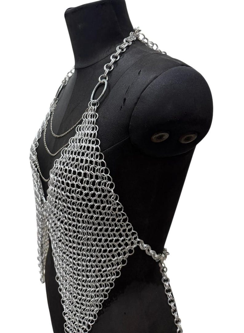 Puede incluir: Top de cota de malla plateada con dise&ntilde;o de cuello halter. La parte superior est&aacute; construida con peque&ntilde;os anillos de metal entrelazados, formando una tela flexible y texturizada. Se muestra en un maniqu&iacute; negro.