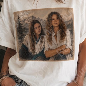 Può includere: Una t-shirt bianca a maniche corte con una stampa grafica rettangolare di due persone con i capelli lunghi sedute davanti a un paesaggio montano. Il testo sterling point è stampato nell'angolo in alto a sinistra dell'illustrazione.