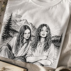 Può includere: Una t-shirt bianca a maniche corte con un'illustrazione grafica in inchiostro nero di due persone sedute davanti a un paesaggio lacustre di montagna. Il testo sterling point è stampato in piccole lettere nere sopra l'illustrazione.