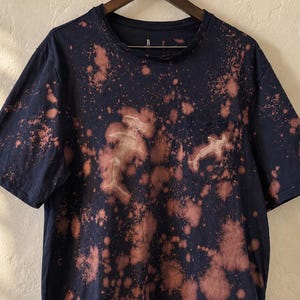 Peut inclure: Un t-shirt bleu marine à manches courtes avec un motif d'éclaboussures décolorées sur tout le tissu. Le vêtement comprend une poche poitrine et deux silhouettes de requins-marteaux dans un ton plus clair et délavé.