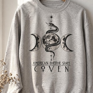 May include: A heather gray crewneck sweatshirt featuring a black graphic print of a snake coiled around a full moon flanked by two crescent moons. Above the moon is a small star. Below the graphic is the text AMERICAN HORROR STORY COVEN.