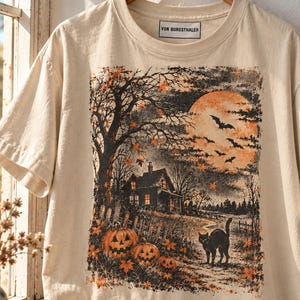 Op de afbeelding: Een crèmekleurig T-shirt met korte mouwen en een grafische print van een spookhuis, een grote oranje maan, vleermuizen, een zwarte kat en drie pompoenen. Het label in de nek bevat de tekst VON BURGSTHALER.