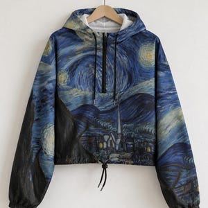 May include: A long-sleeved, cropped pullover hoodie featuring a vibrant, all-over print of a starry night landscape with swirling blue skies, yellow moon-like orbs, and a dark village silhouette. It includes a black half-zip closure and drawstrings.