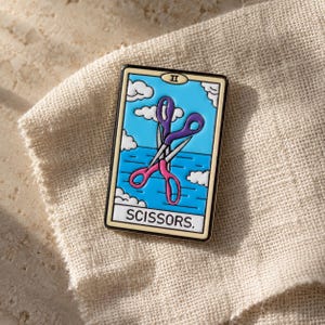 Könnte beinhalten: Eine rechteckige Emaille-Anstecknadel im Design einer Tarotkarte mit hellblauem Hintergrund, weißen Wolken und einer gekreuzten Schere in Lila und Pink. Oben ist die römische Ziffer II zu sehen, unten steht der Text SCISSORS.