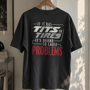 May include: A charcoal gray short-sleeve t-shirt featuring white and red text on the back that reads: IF IT HAS TITS OR TIRES IT'S BOUND TO CAUSE PROBLEMS. The shirt is displayed on a wooden hanger, showcasing a crew neckline and a relaxed fit.