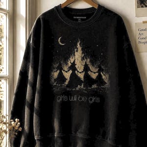 Op de afbeelding: Een zwart sweatshirt met lange mouwen en een grafische print van vier figuren met puntmutsen die elkaars hand vasthouden rond een kampvuur onder een maansikkel. De tekst "girls will be girls" staat in het wit onder de illustratie.