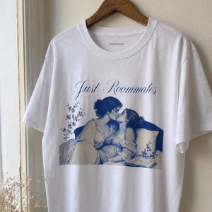 Könnte beinhalten: Ein weißes T-Shirt mit kurzen Ärmeln und einem blauen Grafikdruck von zwei Personen, die sich auf einem Bett umarmen. Der Text "Just Roommates" steht in blauer Schreibschrift über dem Bild. Der Markenname "VON BURGSTHALER" steht im Kragen.