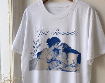May include: A white short-sleeved t-shirt featuring a blue graphic print of two figures embracing on a bed. The text "Just Roommates" is printed in a blue script font above the illustration. The brand name "VON BURGSTHALER" is printed in the collar.