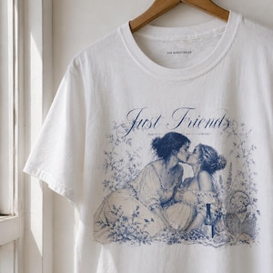 Könnte beinhalten: Ein weißes Kurzarm-T-Shirt mit einem blauen Grafikdruck von zwei Personen, die sich auf einem Feld küssen. Der Text "Just Friends" ist in Schreibschrift über der Illustration gedruckt. Das Shirt hat einen Rundhalsausschnitt und ein "VON BURGSTHALER"-Etikett.