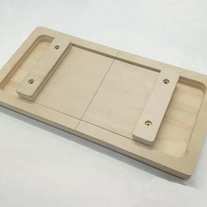 Peut inclure: Un plateau en bois de couleur claire, de forme rectangulaire. Il comporte deux sections surélevées avec des poignées et deux petites pièces en bois fixées avec des vis. Le plateau a une esthétique simple et fonctionnelle.