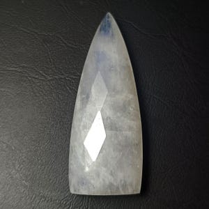 Könnte beinhalten: Ein facettierter, schildförmiger Mondstein-Edelstein mit einem durchscheinenden, milchig-weißen Aussehen und subtilem blauem Schimmer. Der Stein hat eine spitze Oberseite und eine flache Basis, wobei geometrische Facetten das Licht reflektieren.