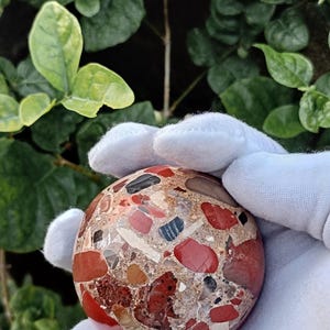 May include: A spherical stone object featuring a polished surface with a conglomerate texture. The sphere displays a mix of embedded fragments in shades of red, brown, black, and cream, set within a light-colored, speckled matrix material.