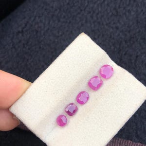 Op de afbeelding: Zes kleine, ronde en vierkant geslepen, levendige roze edelstenen worden getoond op een witte stof. De edelstenen zijn diagonaal gerangschikt en tonen hun gepolijste oppervlakken en rijke kleur. De achtergrond is een donker, getextureerd oppervlak.