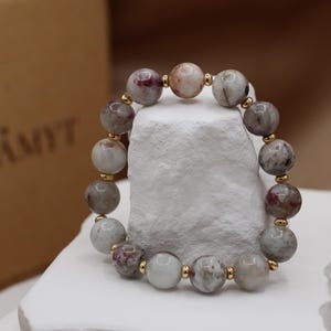 Puede incluir: Una pulsera de cuentas con piedras preciosas grandes y redondas en tonos grises, rosas y blancos, intercaladas con pequeñas cuentas doradas. La pulsera se exhibe sobre una superficie blanca texturizada.