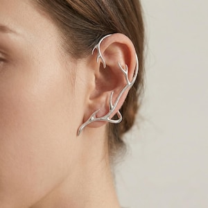 Puede incluir: Un ear cuff plateado con forma de cuernos, que abraza la oreja. La joya presenta un diseño orgánico detallado con múltiples puntas y curvas, creando un accesorio único y elegante.