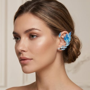 Puede incluir: Un ear cuff plateado con un diseño de pluma esmaltada en azul y blanco. El cuff envuelve la oreja, creando un look único y elegante. El esmalte azul tiene un efecto degradado, añadiendo profundidad al diseño. Esta joya es un accesorio llamativo.