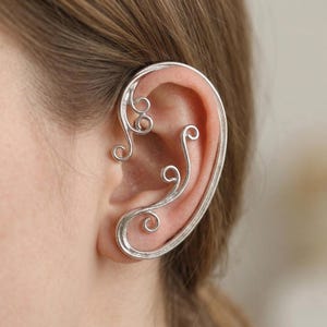 May include: A silver ear cuff with a delicate, swirling design. The ear cuff wraps around the ear, featuring intricate curls and curves. This piece of jewelry is designed to be worn on the ear.