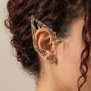 May include: Gold-toned ear cuff featuring a floral and leaf design. The ear wrap is shaped to curve around the ear, with detailed roses and leaves. The jewelry piece is worn on the ear.