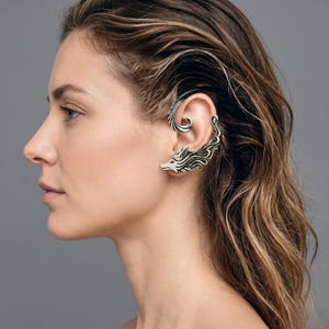 May include: Silver-toned ear cuff featuring a wolf design with a red eye. The cuff wraps around the ear and has a detailed, stylized wolf head. The jewelry is worn on the ear and has a decorative, flowing design.