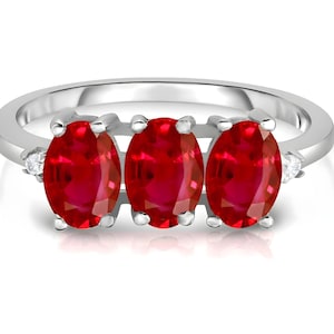 May include: A silver ring featuring three oval-cut, vibrant red gemstones. Small, clear accent stones are set on either side of the central gems. The ring's band is smooth and polished, reflecting light. A piece of jewelry.