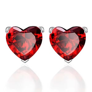 May include: A pair of heart-shaped stud earrings. Each earring features a vibrant red gemstone set in a silver-colored metal. The gemstones are faceted, catching the light and creating a sparkling effect. The earrings are displayed on a white background.