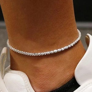 May include: A silver-colored anklet with small, sparkling embellishments. The delicate chain encircles the ankle, reflecting light. The anklet is worn with a white shoe.