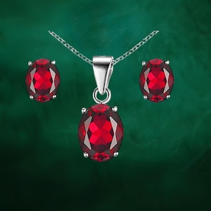 May include: A jewelry set featuring a pendant necklace and matching earrings. The set includes a silver chain necklace with a large, oval-cut, deep red gemstone pendant. The earrings also feature oval-cut red gemstones. The gemstones are set in silver metal.