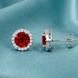 May include: A pair of stud earrings featuring a vibrant red gemstone surrounded by a halo of smaller, clear stones. The earrings have a silver-colored metal setting and posts, set against a teal-colored background.