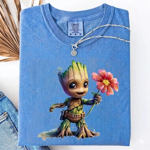 Puede incluir: Una camiseta Comfort Colors azul claro con un personaje de dibujos animados que sostiene una flor rosa. El personaje es un humanoide parecido a un árbol con un cinturón marrón y una pequeña bolsa. Un collar plateado con un colgante en forma de sol está sobre la camiseta.
