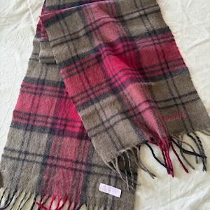 May include: A rectangular scarf featuring a plaid pattern in shades of olive green, dark navy blue, and vibrant magenta. The scarf has a soft, textured appearance with fringed ends and a small rectangular white label attached near one of the edges.