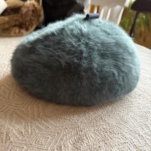 May include: A teal-colored, fluffy beret hat with a small, dark blue accent. The hat is round and sits on a patterned, off-white tablecloth. Other hats are visible in the background.