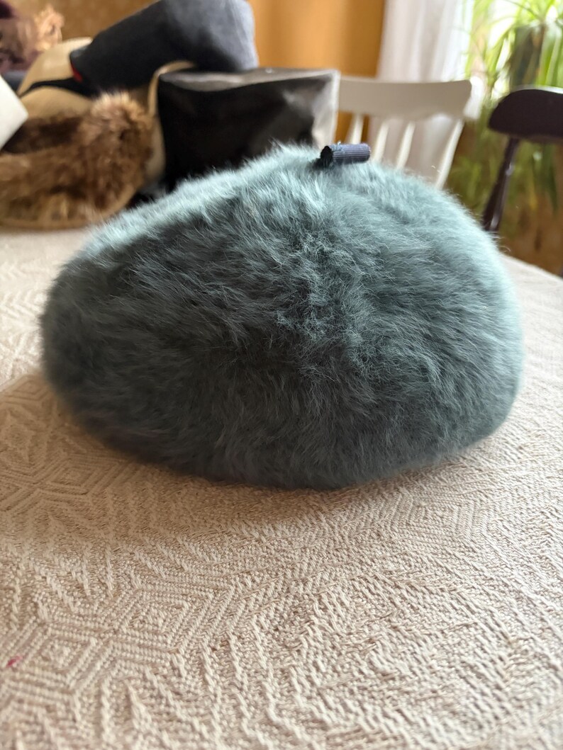 May include: A teal-colored, fluffy beret hat with a small, dark blue accent. The hat is round and sits on a patterned, off-white tablecloth. Other hats are visible in the background.