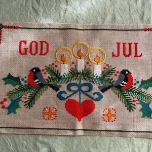 以下が含まれることがあります： ベージュの布地に赤い文字で「GOD JUL」と刺繍された長方形のクロスステッチ。黄色い炎の白いキャウドル3本、ウソの鳥2羽、緑の松の枝、ヒイラギの葉、赤い実、そして赤いハートがデザインされています。