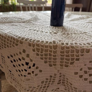 May include: A white, crocheted tablecloth with a detailed openwork pattern covers a table. A dark blue vase holding a green plant sits on the table. The tablecloth's edge is visible, showing the intricate design.