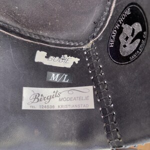 May include: Close-up of a black leather hat with white stitching and labels. The labels read "BUGIE", "M/L", and "Birgits Modeatelje TEL. 124536 KRISTIANSTAD". A circular sticker reads "HEAD 'N HOME SANTA CRUZ CA".