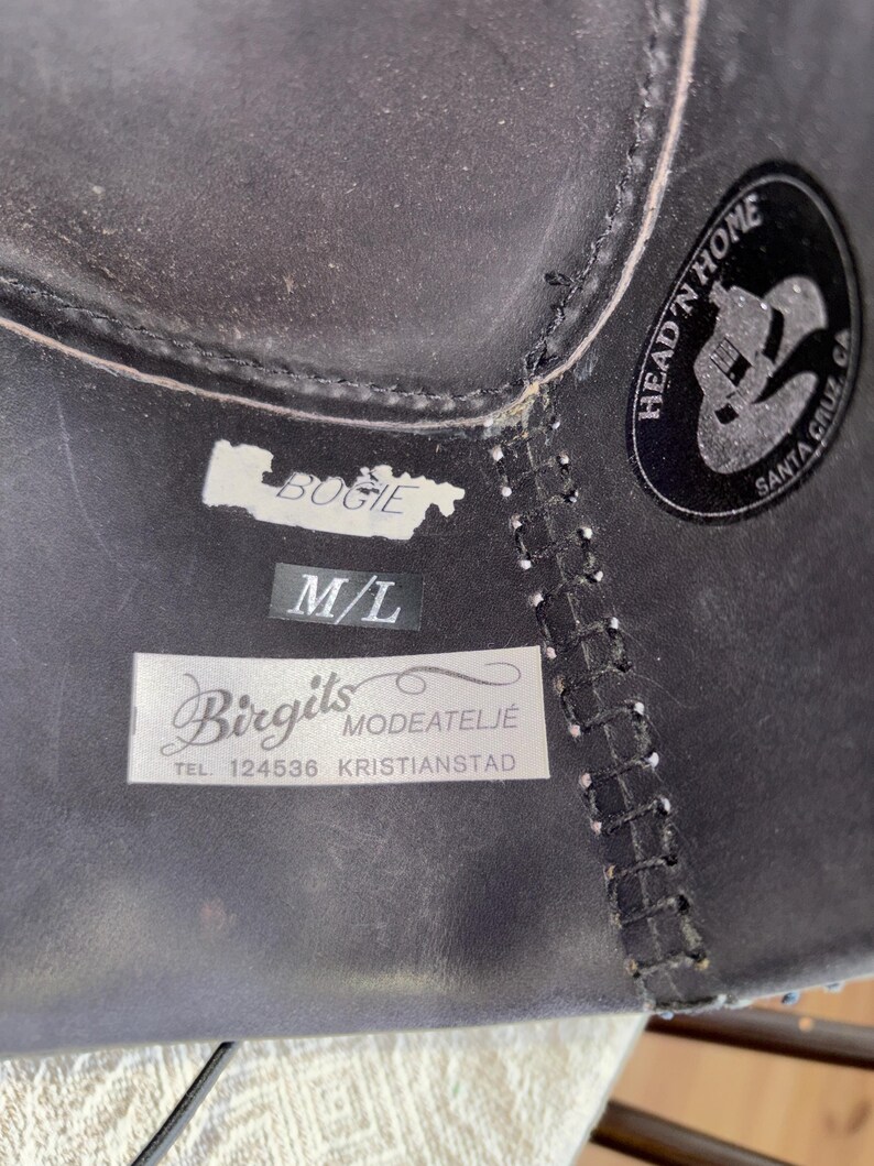 May include: Close-up of a black leather hat with white stitching and labels. The labels read "BUGIE", "M/L", and "Birgits Modeatelje TEL. 124536 KRISTIANSTAD". A circular sticker reads "HEAD 'N HOME SANTA CRUZ CA".