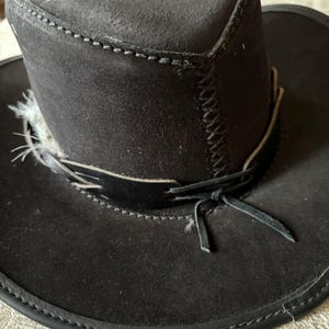 May include: A black suede cowboy hat with a wide brim and a decorative band. The hat features a stitched design around the crown and brim, and a feather accent. The hat has a leather band tied in a bow.