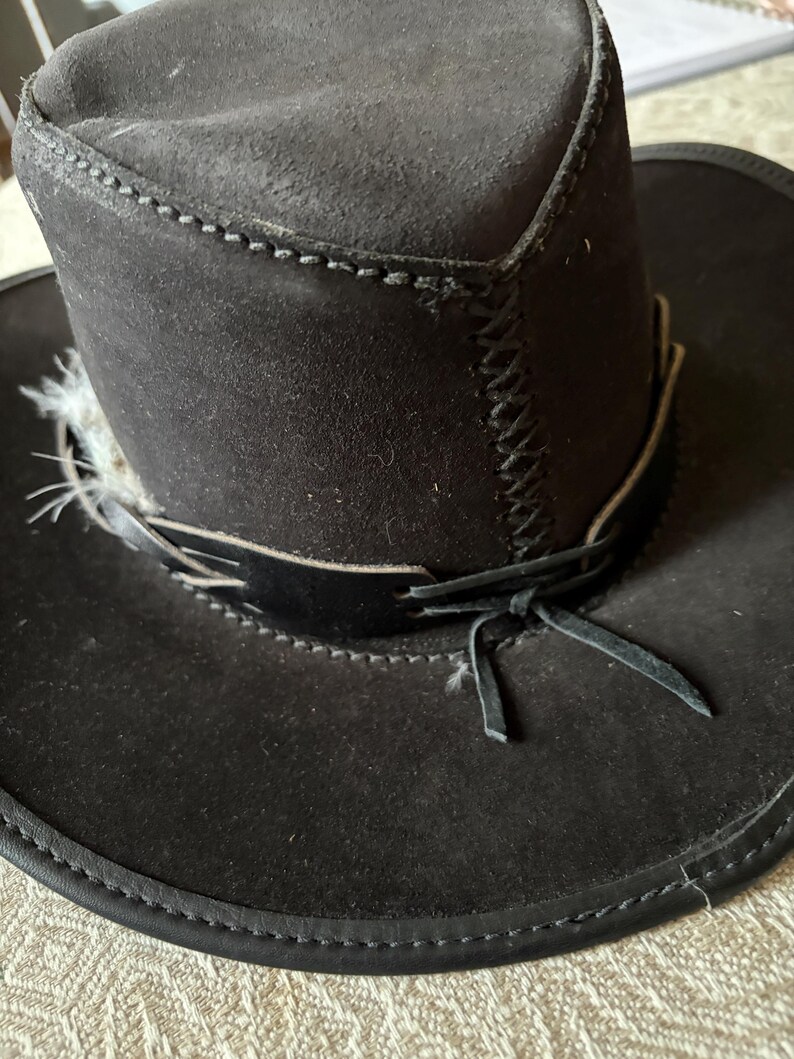 May include: A black suede cowboy hat with a wide brim and a decorative band. The hat features a stitched design around the crown and brim, and a feather accent. The hat has a leather band tied in a bow.