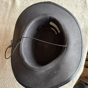 May include: A dark gray cowboy hat with a black leather brim and a black leather chin strap. The hat has a stitched design around the brim and a label inside. The hat is sitting on a patterned surface.