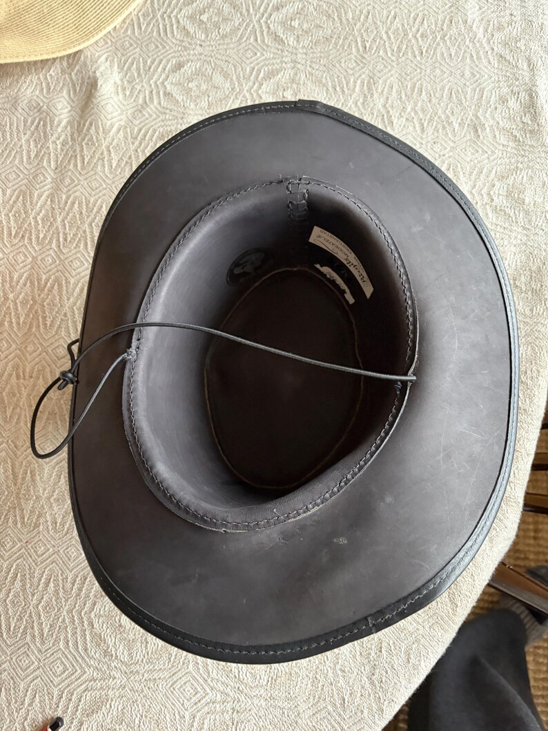 May include: A dark gray cowboy hat with a black leather brim and a black leather chin strap. The hat has a stitched design around the brim and a label inside. The hat is sitting on a patterned surface.