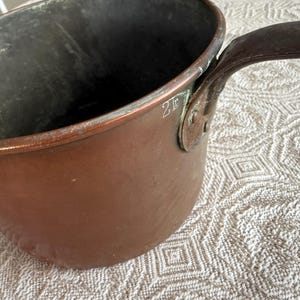 May include: A copper saucepan with a dark interior and a riveted handle. The pot has a rounded shape and a slightly flared rim. The number "2f" is etched on the side. The handle is made of a darker metal.