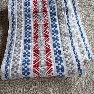 May include: White woven textile with a geometric pattern in red, blue, and grey. The design features a central red band with diamond shapes, flanked by blue and grey floral-like motifs. The edges are trimmed with blue.