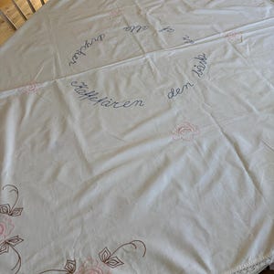 Könnte beinhalten: Ein runder, cremefarbener Tischläufer mit gestickten rosa Rosen und braunen Stielen. Der Tischläufer hat einen Text in Schreibschrift. Der Rand des Tischläufers hat eine zarte Spitzenborte.